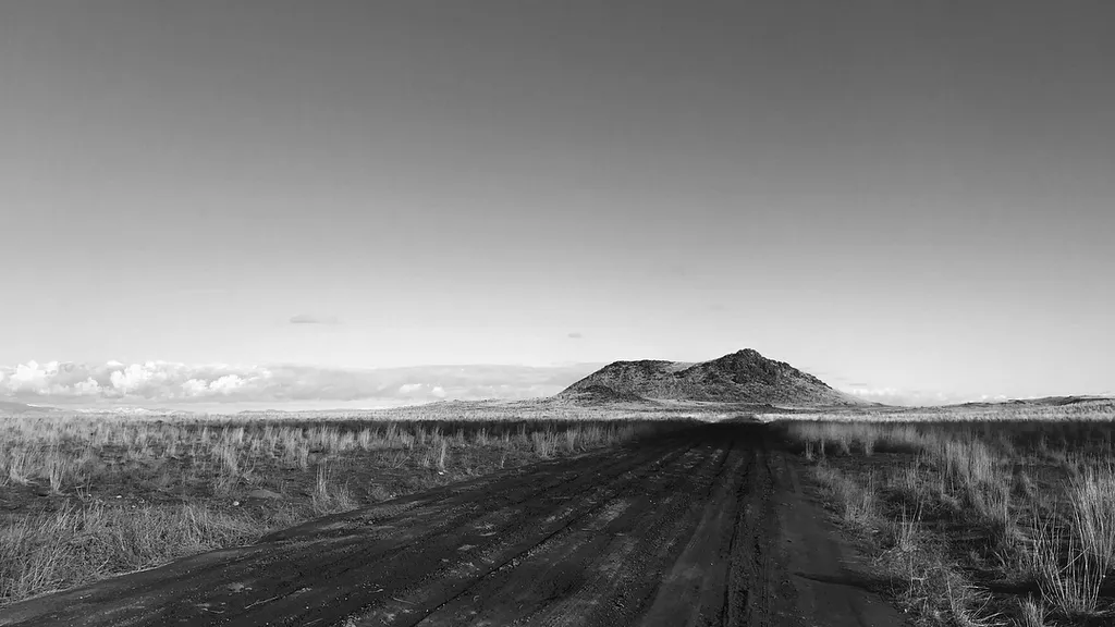 A monochrome landscape showing a dark, straight road cutting through tall grasses toward a solitary, rounded hill under a bright, expansive sky.