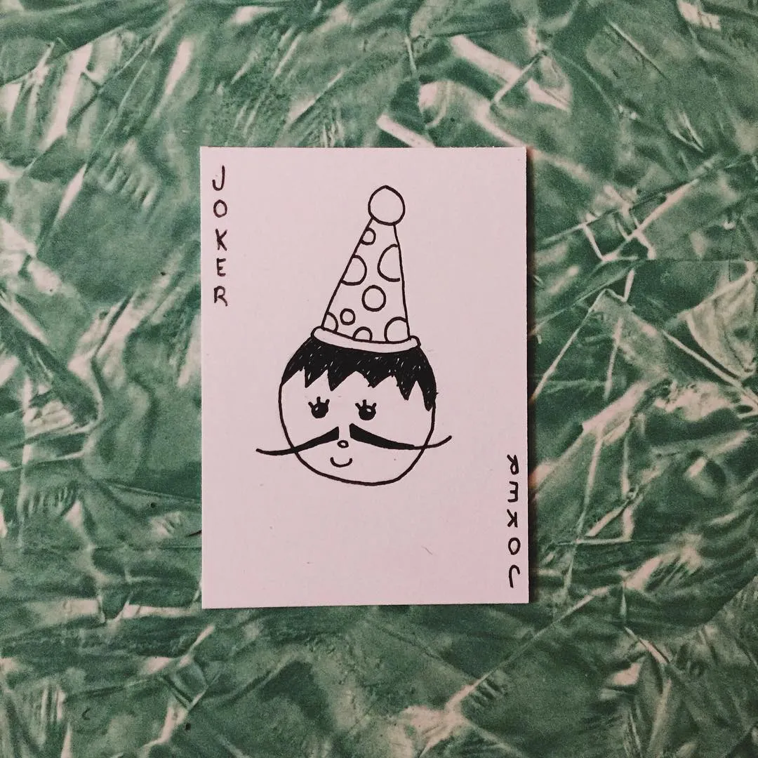 A white card features a black line drawing of a cartoon joker with a spotted hat, flanked by the word JOKER, set against a mottled green and white background.