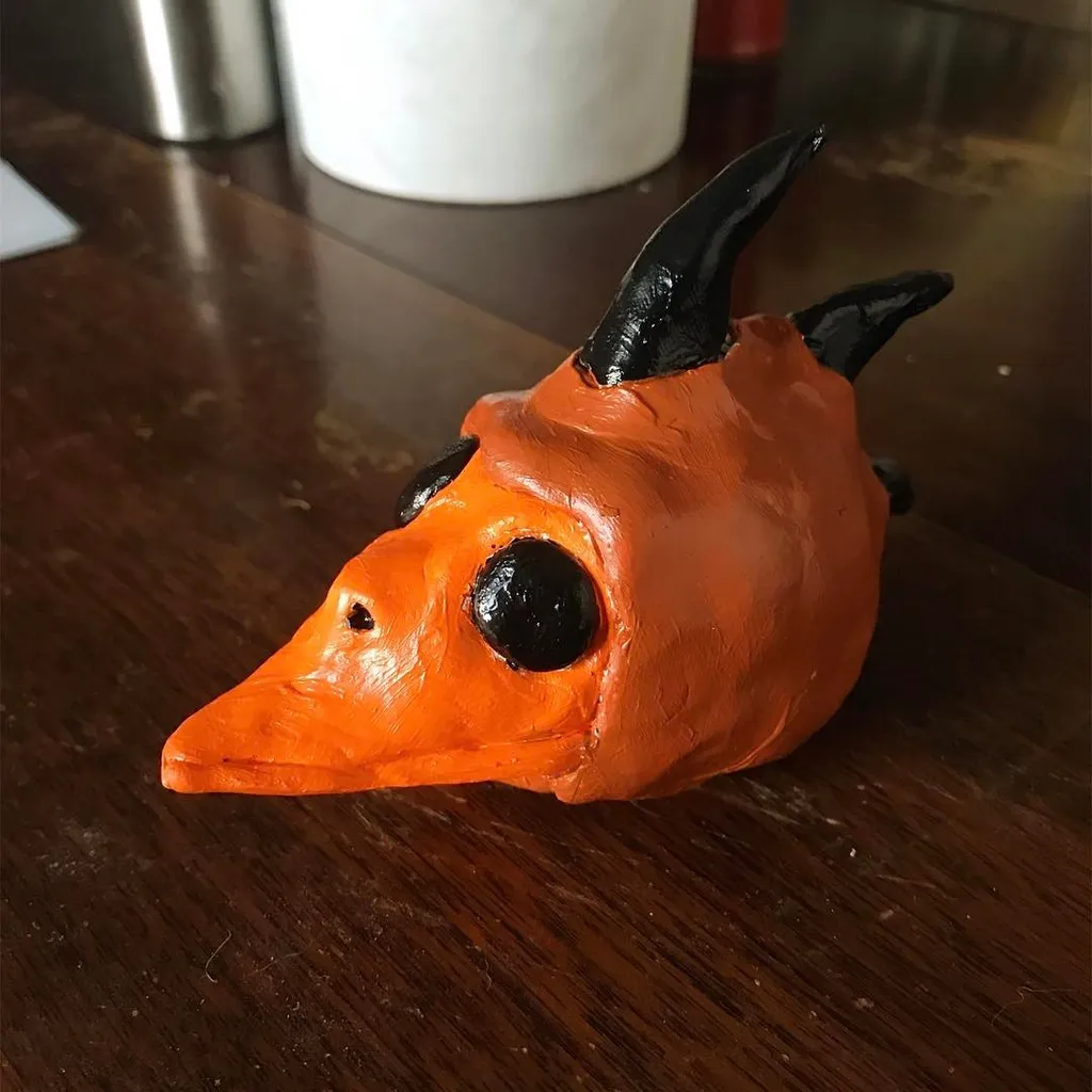A stylized orange sculpture of a fox head, featuring black eyes and two curved black horns, sits on a dark, polished wooden surface.
