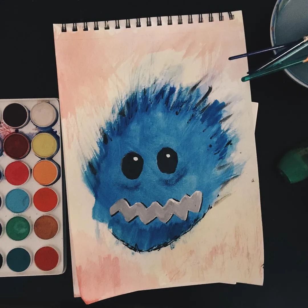 A bright blue, round creature with large black eyes and a jagged mouth is drawn on lined paper that has soft pink and orange washes around the edges.