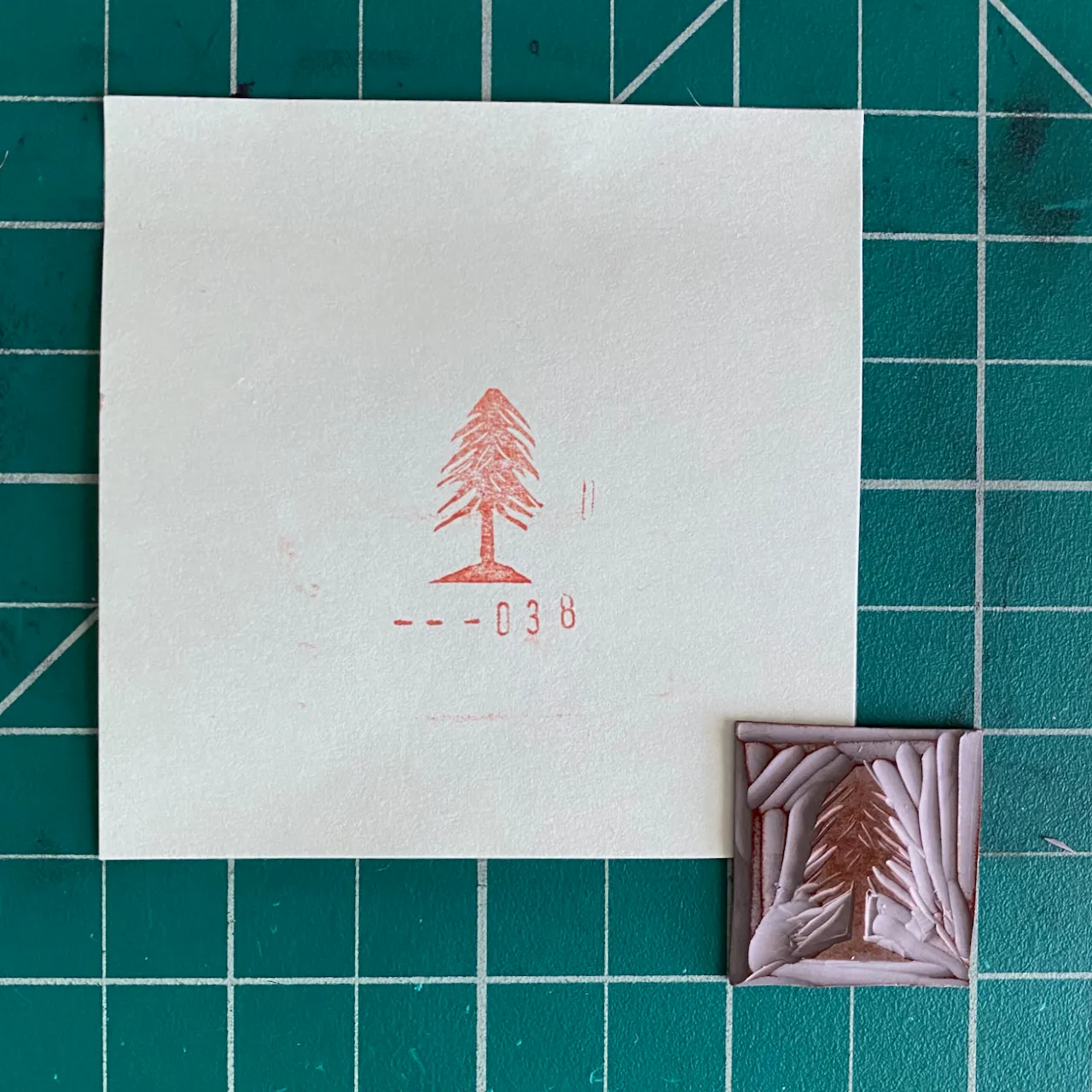 A white card featuring a reddish pine tree drawing and the number --038 is positioned next to a small, square plaque with a raised, carved pine tree design in muted pinks and browns, all set on a green grid.