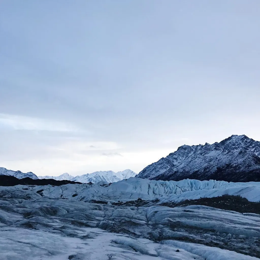 Vast, cool landscape under a pale gray sky, featuring rippled icy ground in the foreground, leading to rugged, snow-dusted mountains and glacial blue formations.