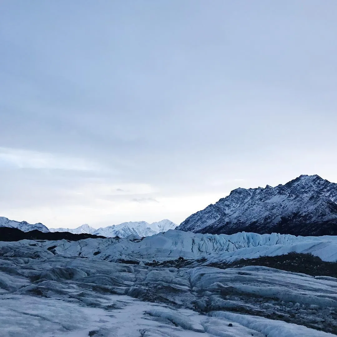 Vast, cool landscape under a pale gray sky, featuring rippled icy ground in the foreground, leading to rugged, snow-dusted mountains and glacial blue formations.