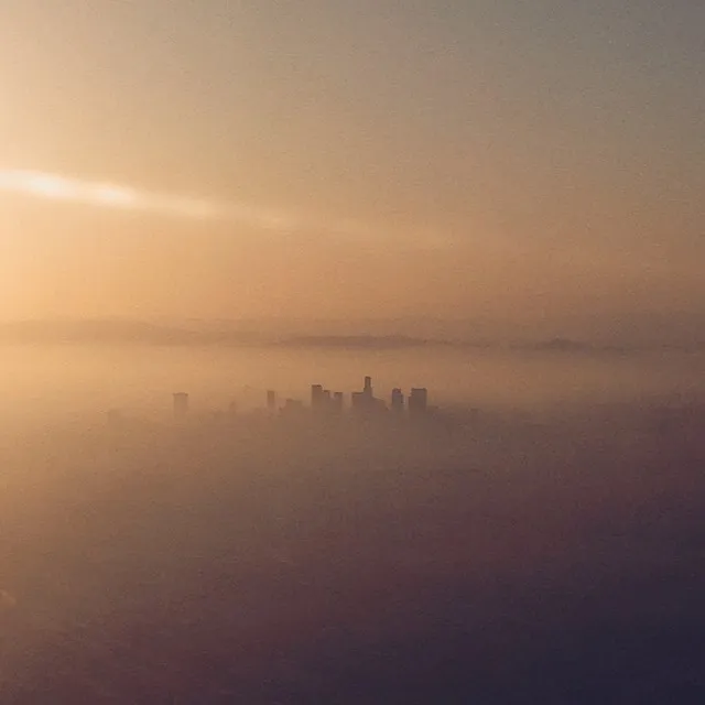 Low-lying skyscrapers are silhouetted in a dense, warm fog, framed by a gradient sky of soft gold and pale orange.