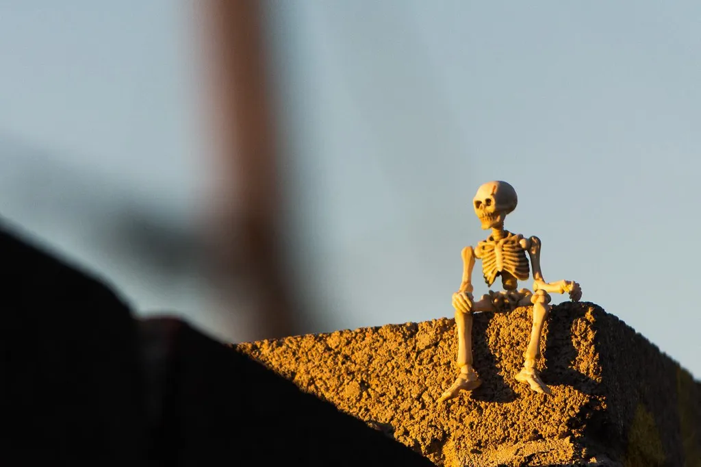 A small skeleton sits on a mound of granular yellow material, silhouetted against a bright, hazy sky with a dark foreground element.