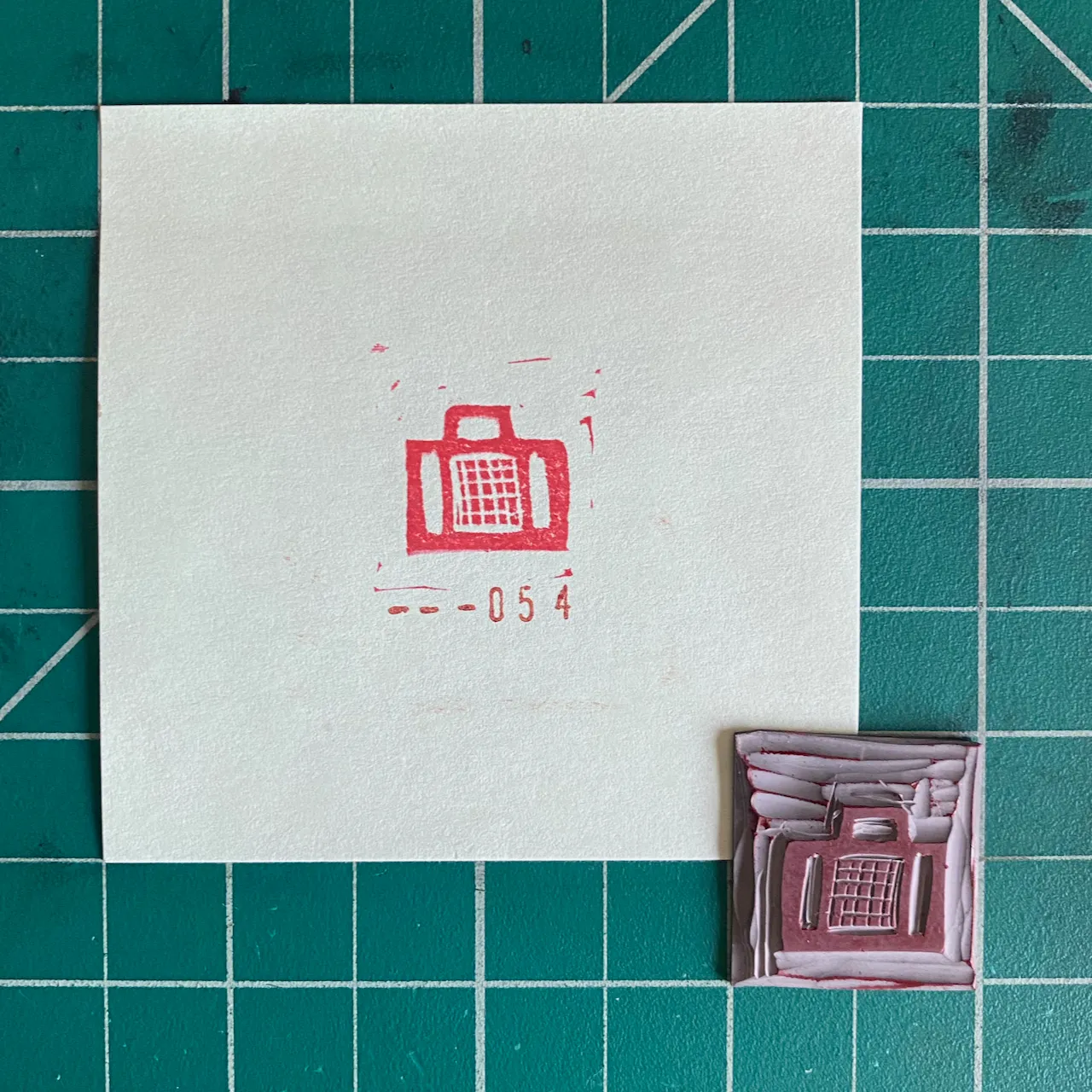 A white card on a teal grid features a red drawing of a suitcase and the number -054, beside a small, textured, reddish-brown piece with a raised red suitcase design.