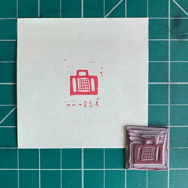 A white card on a teal grid features a red drawing of a suitcase and the number -054, beside a small, textured, reddish-brown piece with a raised red suitcase design.