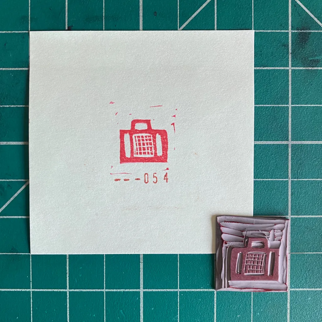A white card on a teal grid features a red drawing of a suitcase and the number -054, beside a small, textured, reddish-brown piece with a raised red suitcase design.