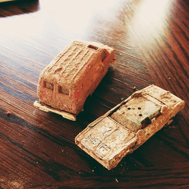 Two distressed, reddish-brown miniature vehicles rest on a dark, polished wooden surface, showing visible grain and strong reflections from overhead light.