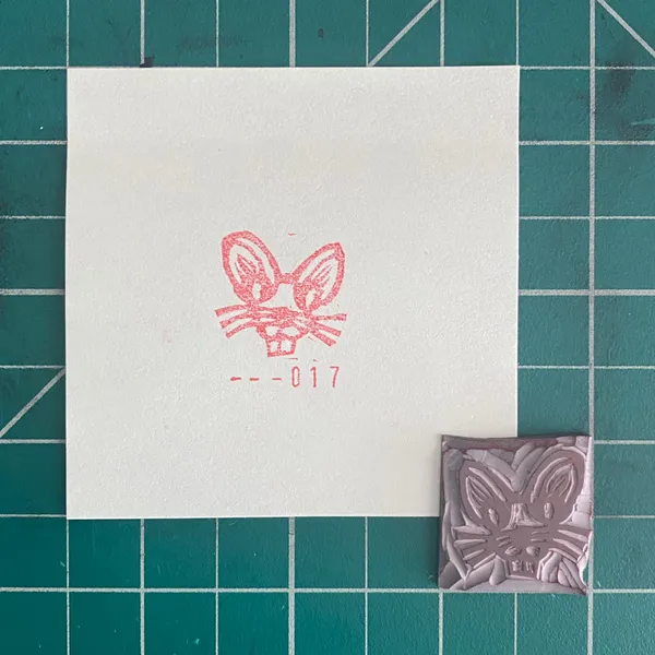 A white card on a teal grid displays a pink outline drawing of a rabbit face with the markings --017, next to a small, square, silver embossed rabbit design.