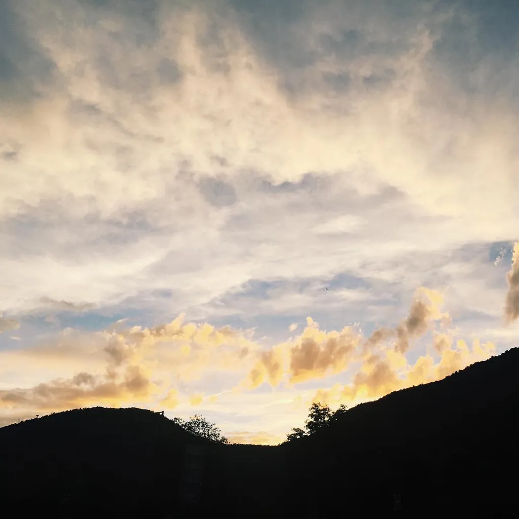 Dark silhouetted hills frame a valley beneath a vast sky filled with layered, creamy yellow and gray clouds illuminated by soft golden light near the horizon.