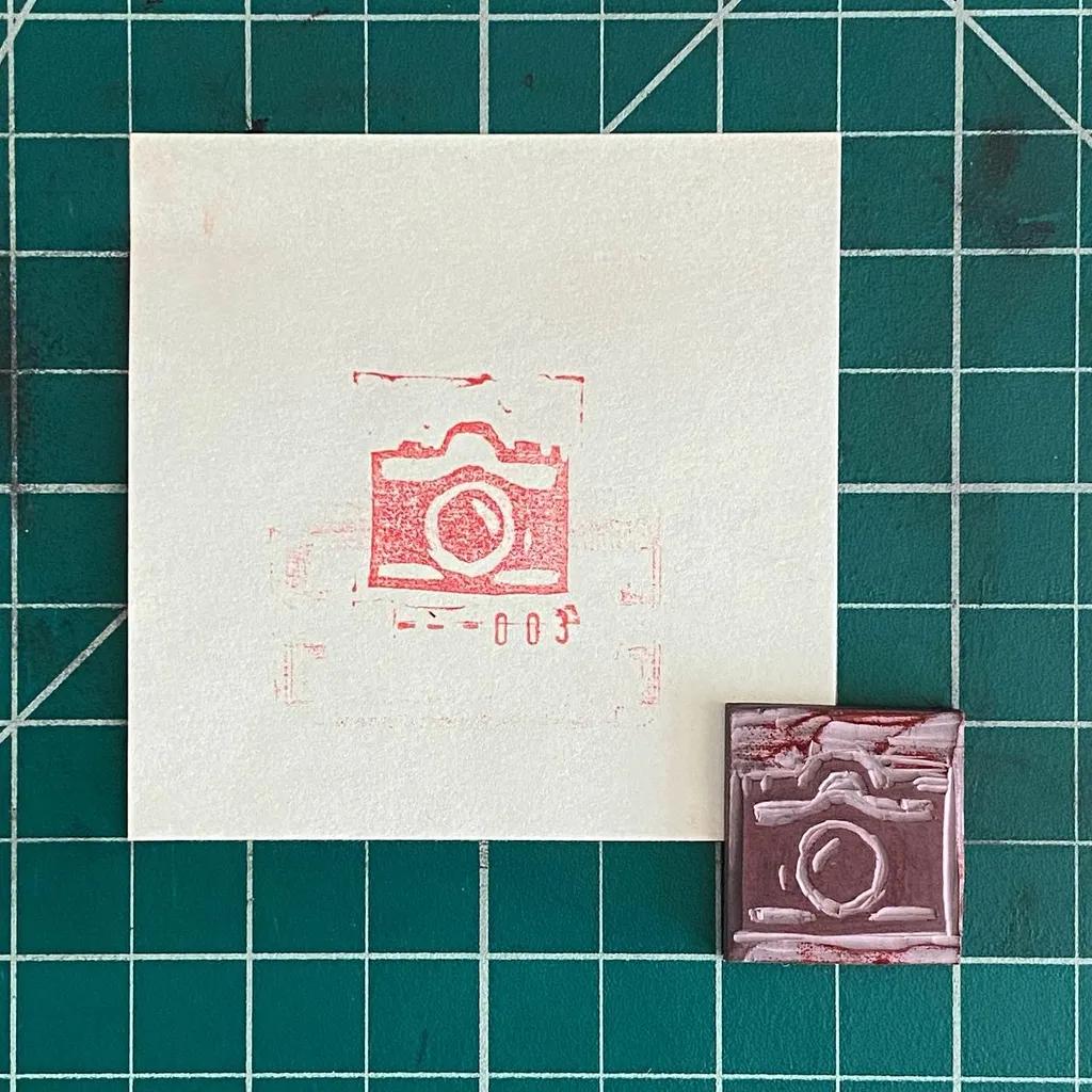 A teal grid background holds a large off-white square with a red stamped camera design, positioned above a smaller reddish-brown rectangular piece also bearing a stamped camera design.
