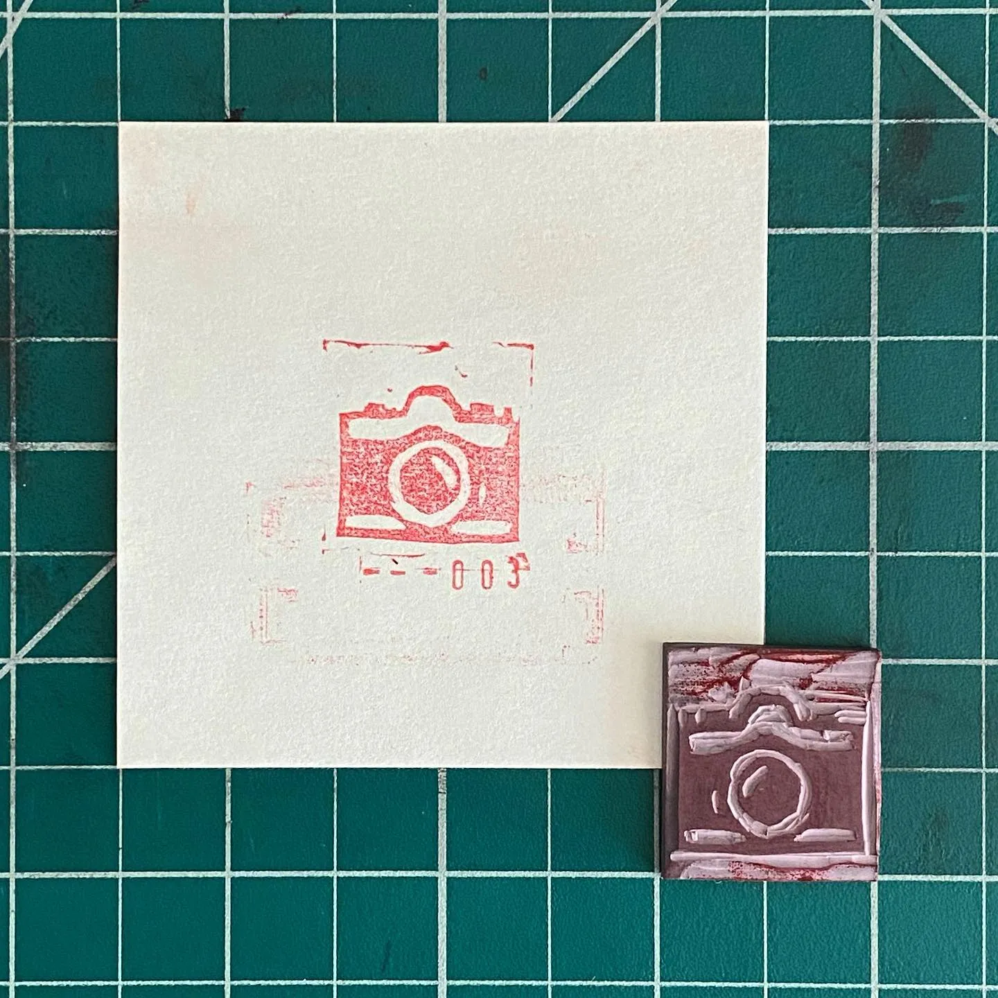 A teal grid background holds a large off-white square with a red stamped camera design, positioned above a smaller reddish-brown rectangular piece also bearing a stamped camera design.