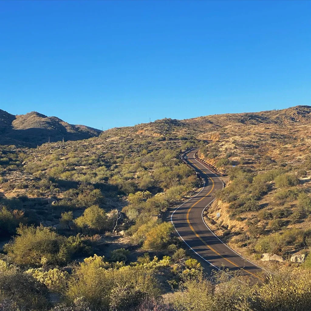 A winding asphalt road traverses arid, scrub-covered hills under a vast, clear blue sky, featuring earth tones and deep greens.