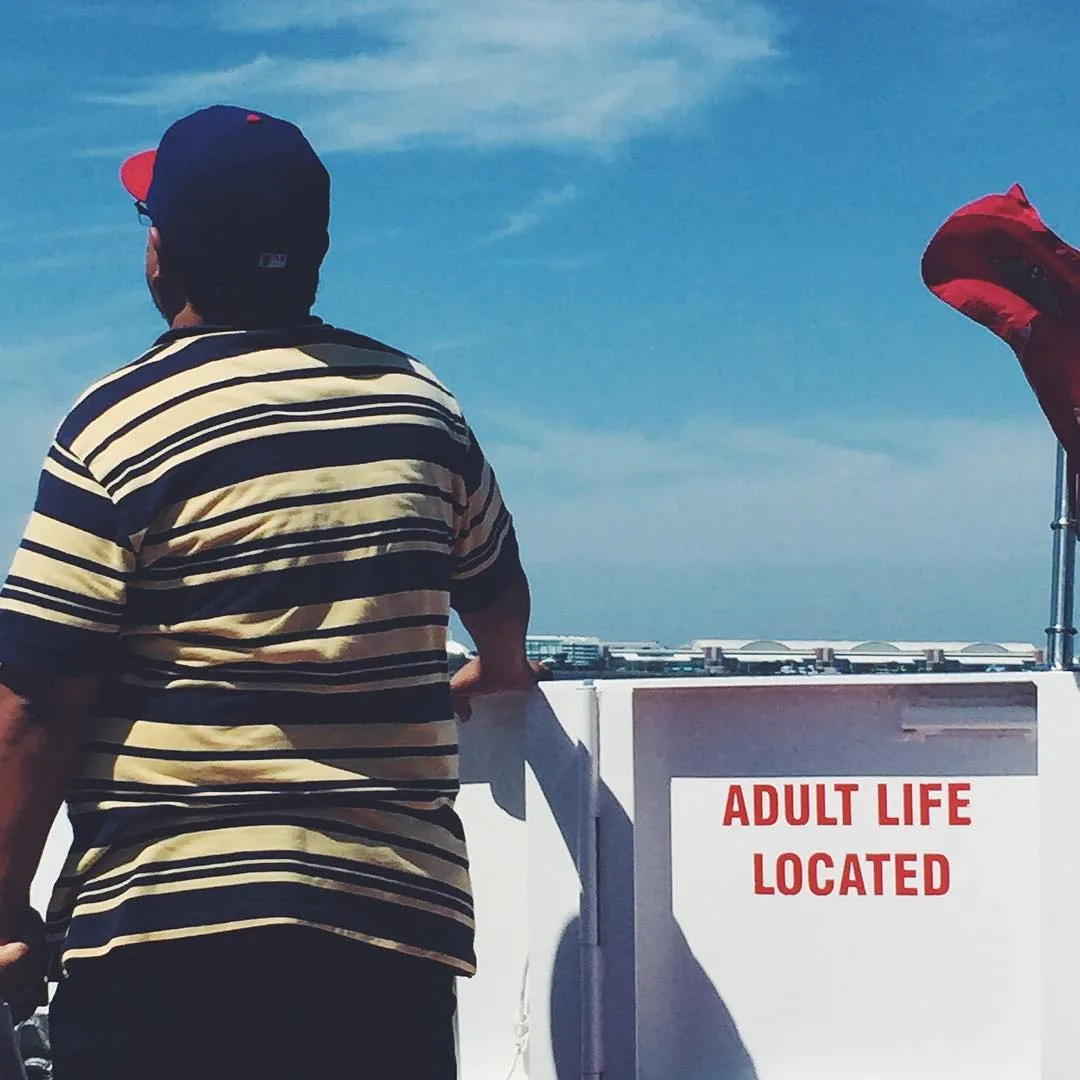 A man in a striped shirt and dark cap stands on a white platform under a bright blue sky, facing red lettering that reads ADULT LIFE LOCATED.