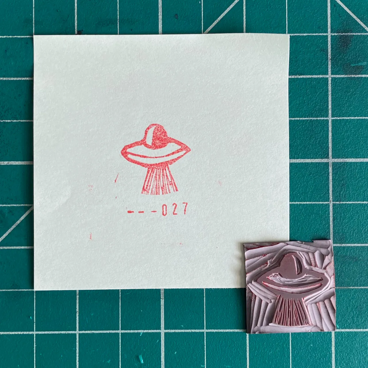 A white paper displays a red ink drawing of a flying saucer and the number --027, positioned next to a square, textured block showing a raised, pink and purple impression of the same saucer.