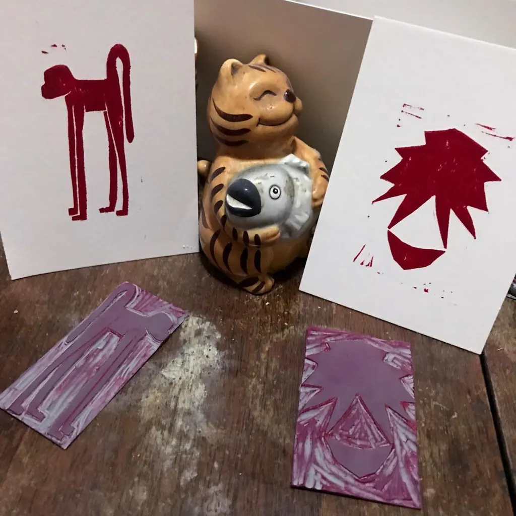 A striped cat figurine sits on a wooden surface surrounded by papers displaying red stick figures, abstract red shapes, and two rectangular pieces marked with pink and purple geometric patterns.