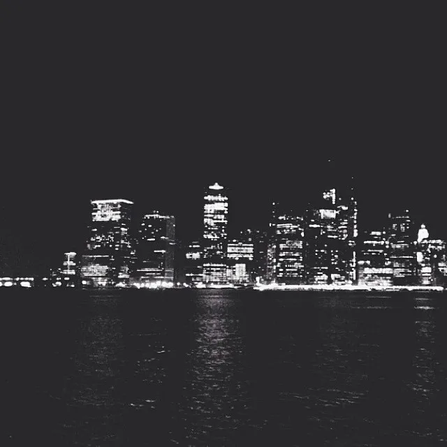 A high-contrast nighttime view shows a dense, illuminated city skyline across dark water, featuring bright yellow and white lights against deep black tones.