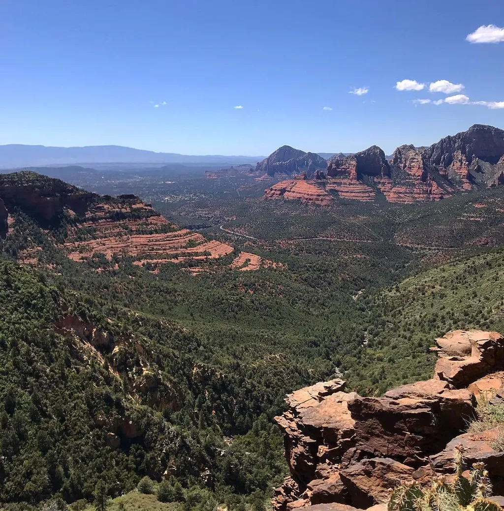 Foreground rocks frame a sweeping vista of deep green scrubland and layered reddish-brown mesas, meeting a hazy blue mountain range under a bright blue sky.