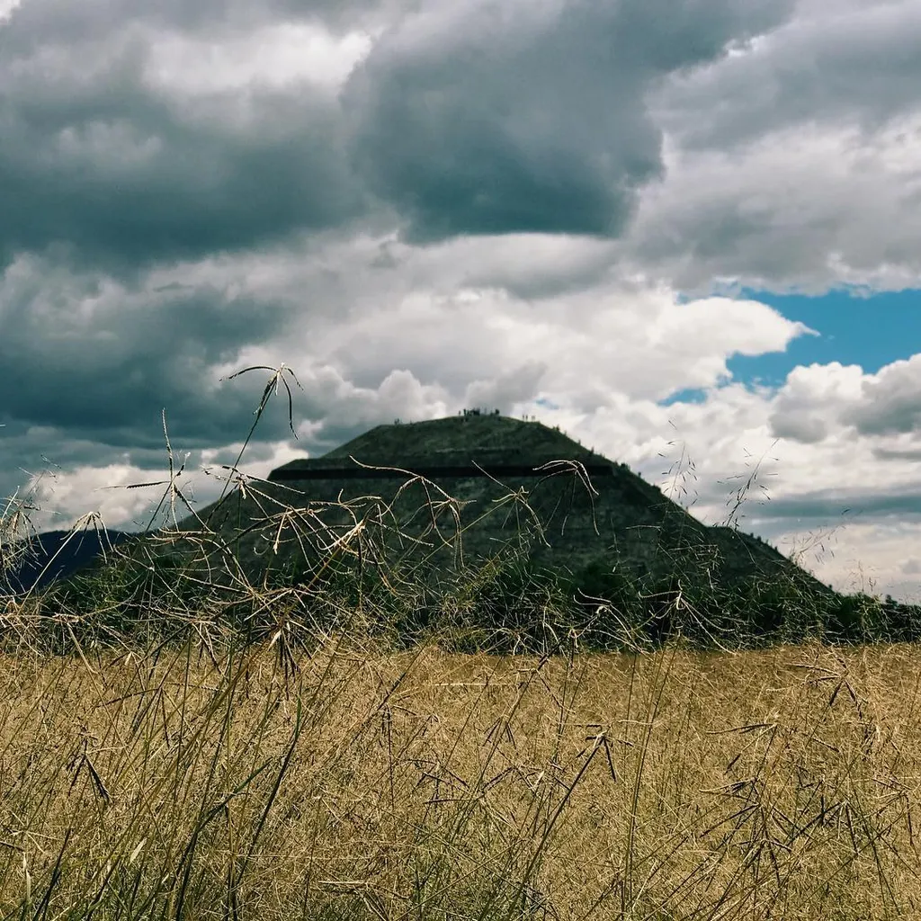 Dry golden grasses fill the foreground below a dark, conical hill set against a dramatic sky of heavy gray clouds and patches of bright blue.