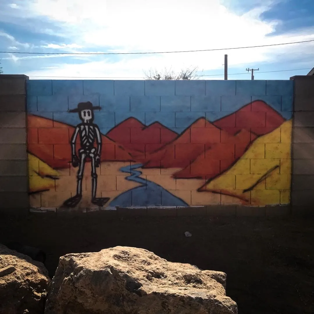 A mural painted on a wall features a stylized skeleton figure wearing a hat, standing before a landscape of layered red, orange, and yellow mountains with a blue river winding through them.