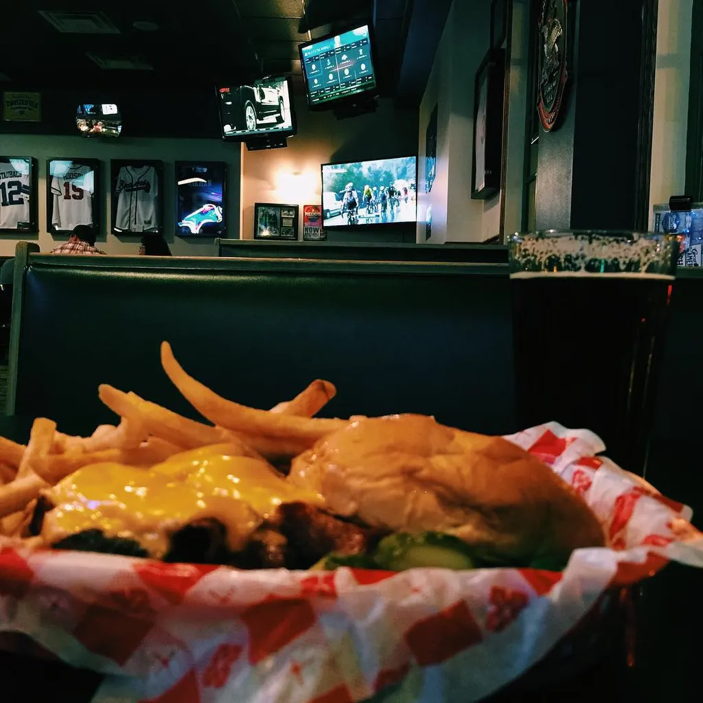 A plate of cheesy fries, meat, and a bun rests on a red and white checkered liner, set against a dark background featuring multiple glowing digital screens and restaurant seating.