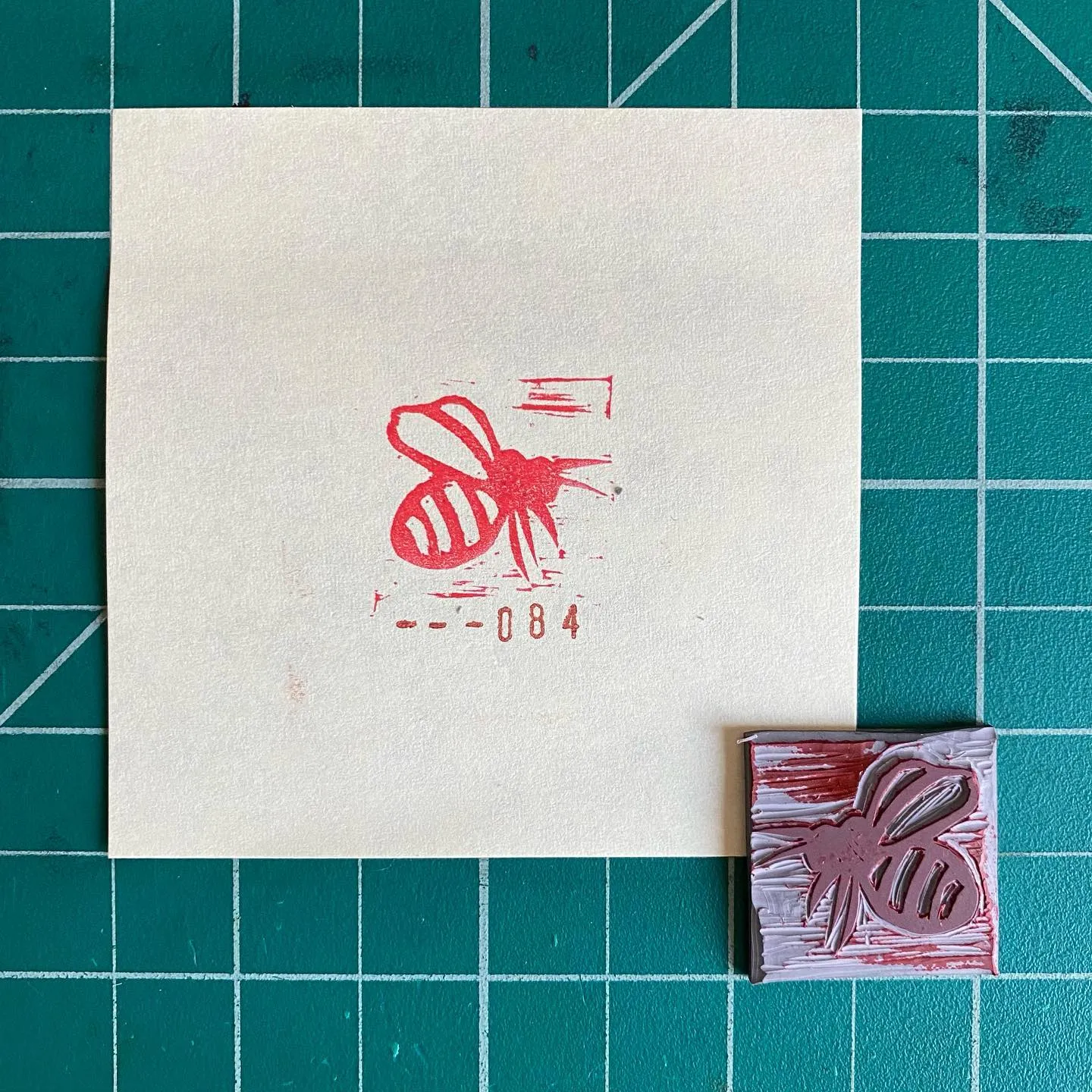 On a teal grid, a cream card displays a red stylized bee and the number 1-084, positioned next to a small square block with a red bee design and visible white texture marks.