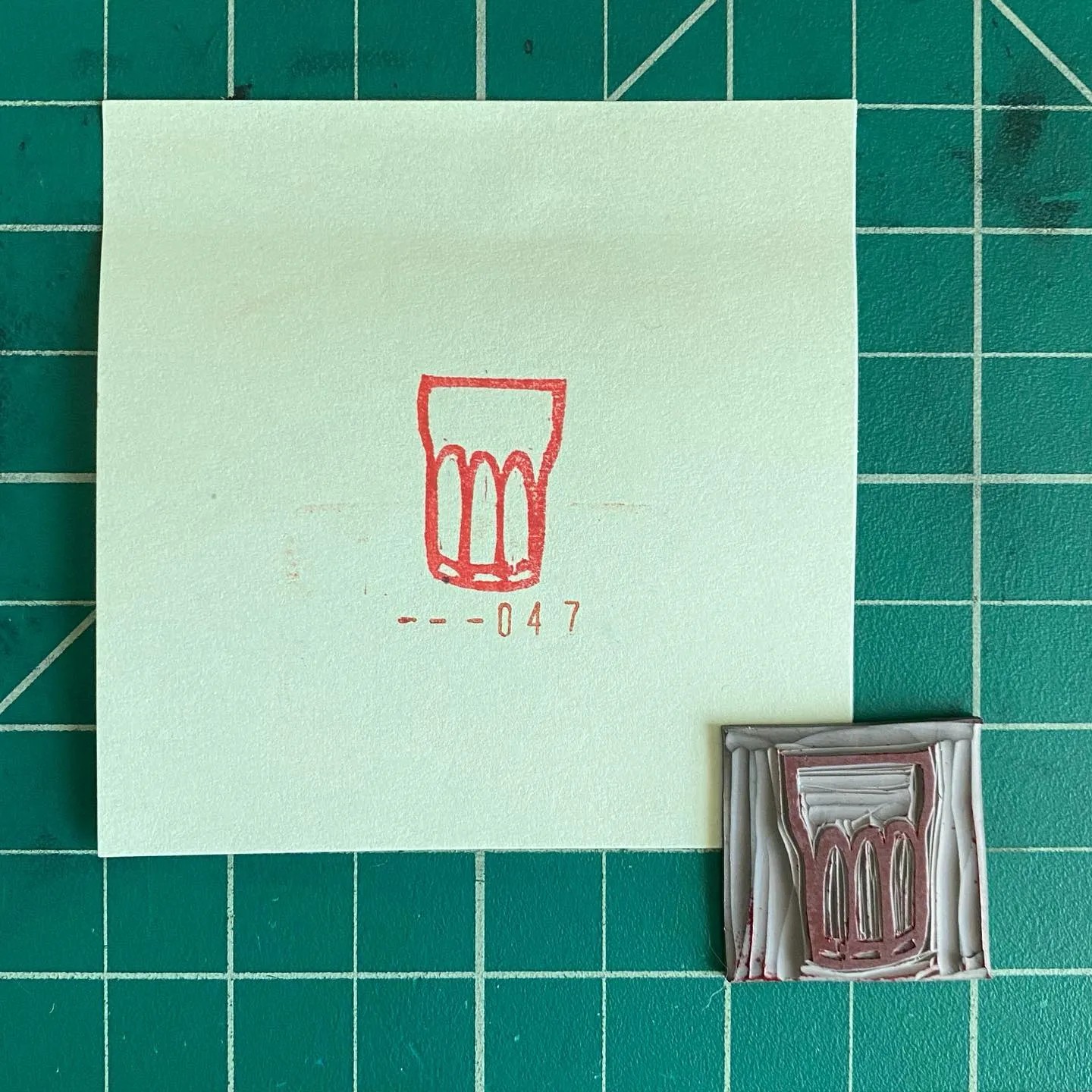 A pale square paper on a dark green grid displays a red outline drawing of a glass and the number --047, next to a small, rectangular, layered object in brown and gray tones.