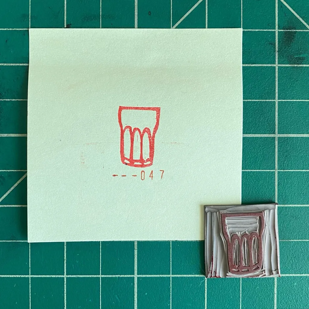 A pale square paper on a dark green grid displays a red outline drawing of a glass and the number --047, next to a small, rectangular, layered object in brown and gray tones.