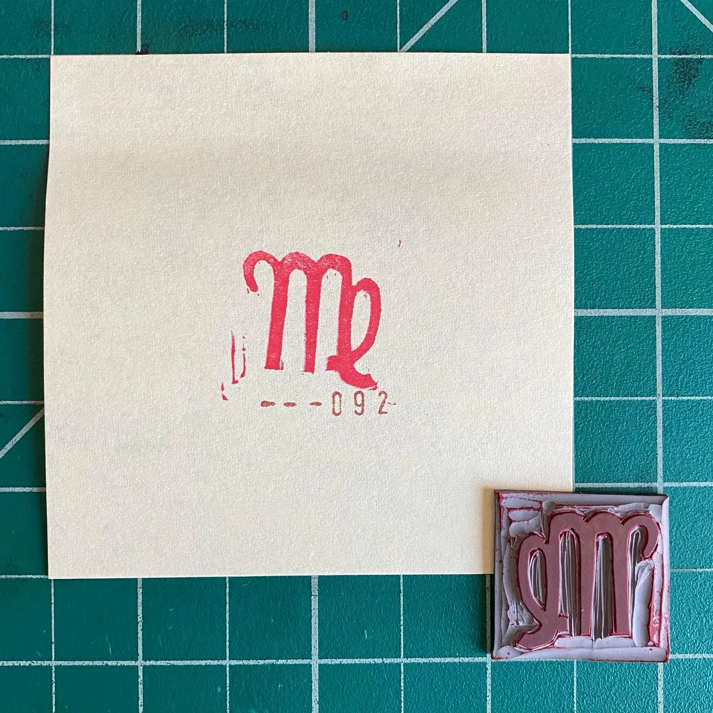 A cream square paper on a teal grid displays a red monogram MR and the numbers 092, with two smaller, painted squares featuring the same red monogram placed below.