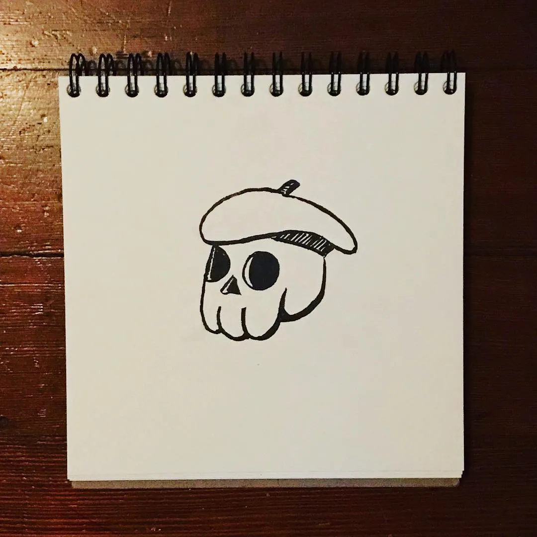 A simple black line drawing of a cartoon skull wearing a small hat, centered on a white spiral notepad resting on dark wood.