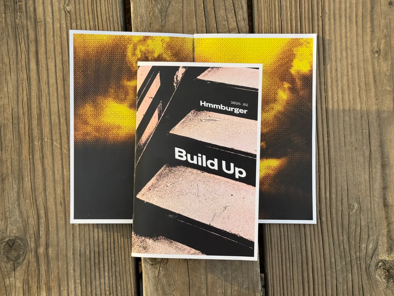 Layered flyers featuring black and yellow geometric patterns rest on a weathered wooden surface, with the central flyer displaying the text Build Up and Hamburger over distressed brown textures.