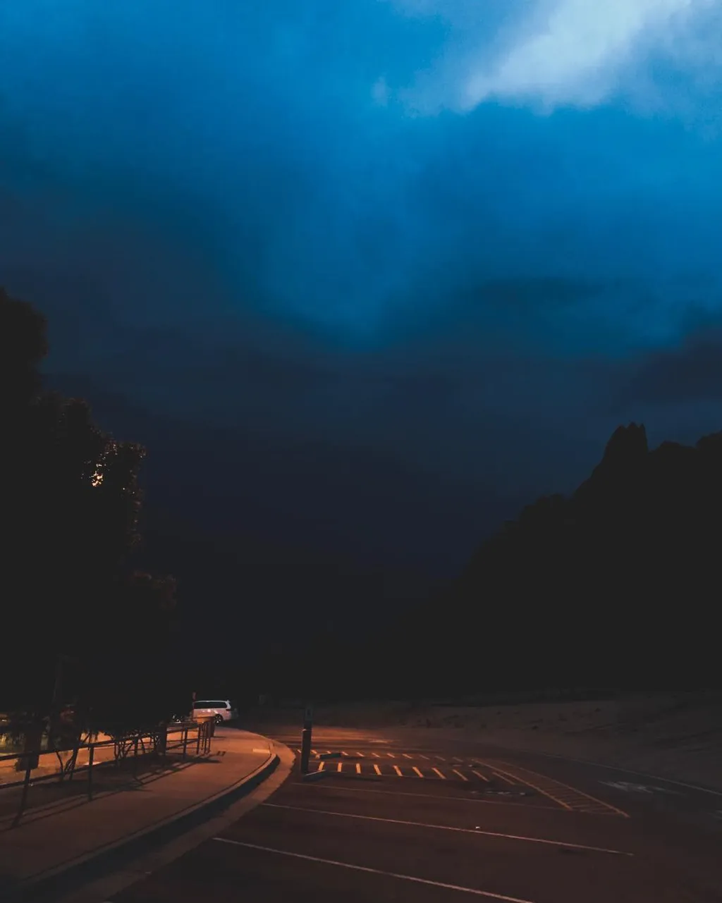 Deep blue and teal clouds dominate the sky above a dark, paved foreground showing parking lines and a silhouetted mountain range on the right.