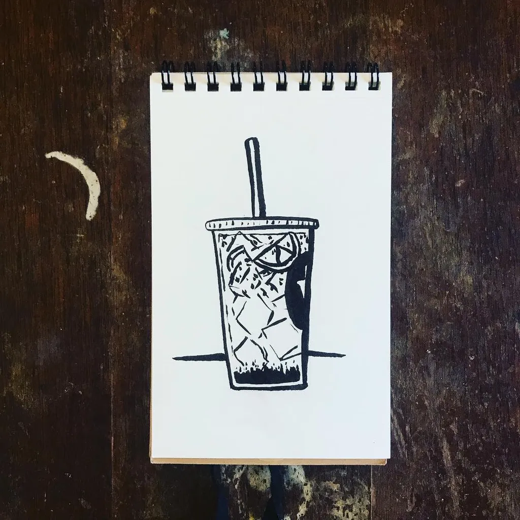 Black ink sketch of a tall, iced beverage in a cup, featuring condensation and a straw, drawn on a white spiral notebook page resting on dark, textured wood.