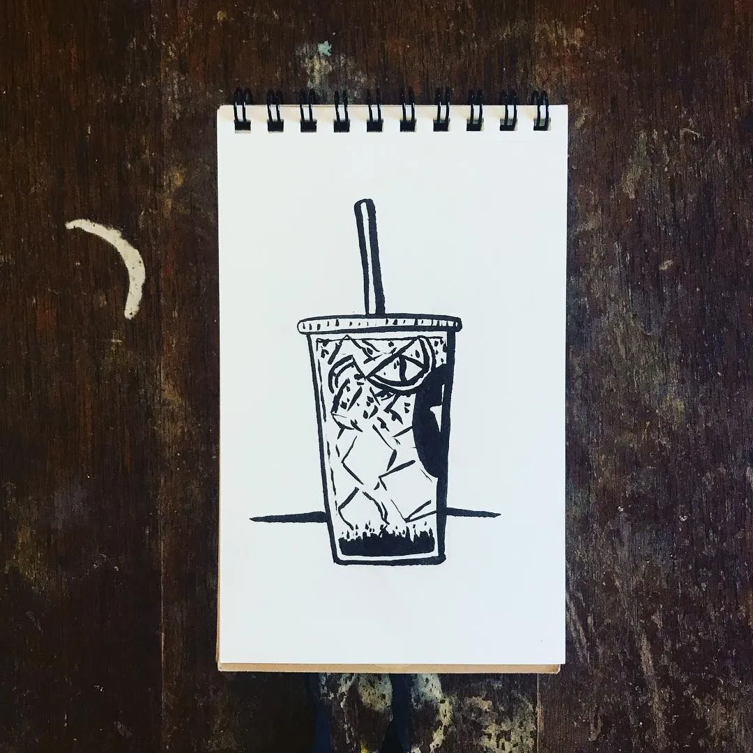 Black ink sketch of a tall, iced beverage in a cup, featuring condensation and a straw, drawn on a white spiral notebook page resting on dark, textured wood.