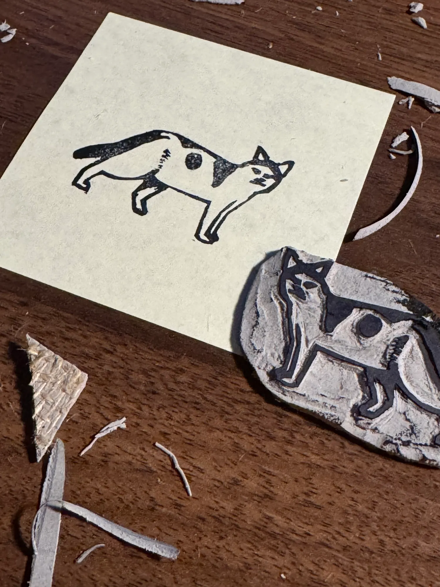 On a dark wood surface, a cream paper sheet holds a black ink drawing of a cat, positioned near a textured plaque displaying a black and white drawing of a dog.