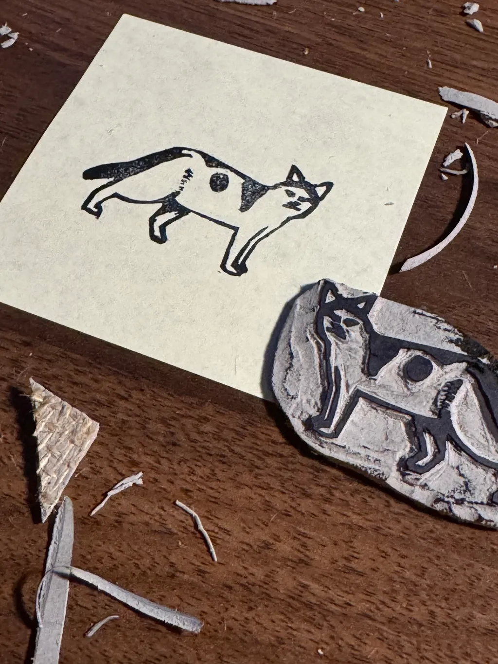 On a dark wood surface, a cream paper sheet holds a black ink drawing of a cat, positioned near a textured plaque displaying a black and white drawing of a dog.
