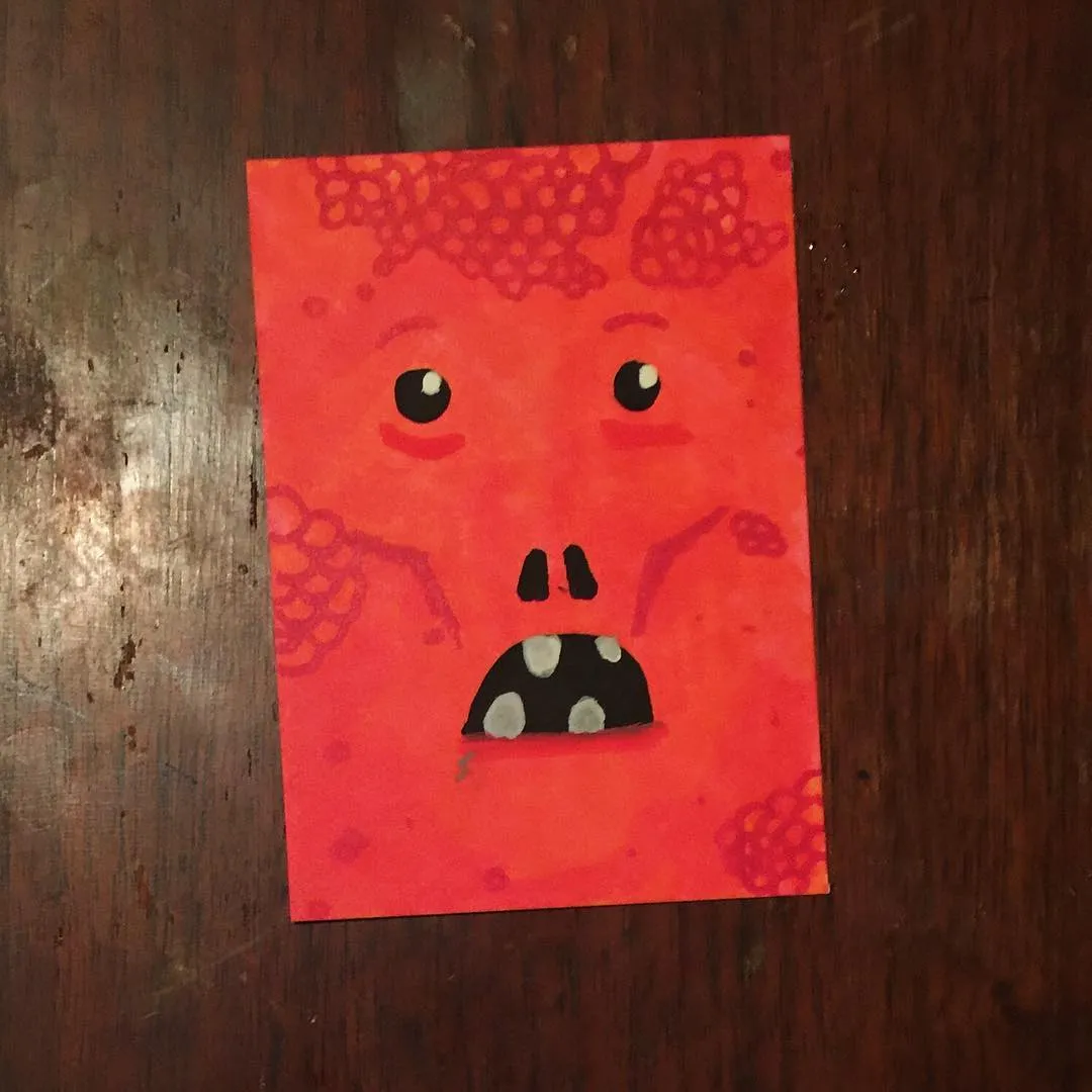 A stylized red face with black eyes, a black nose, and a mouth showing white teeth is centered on a square piece of red paper marked with irregular darker red patterns.