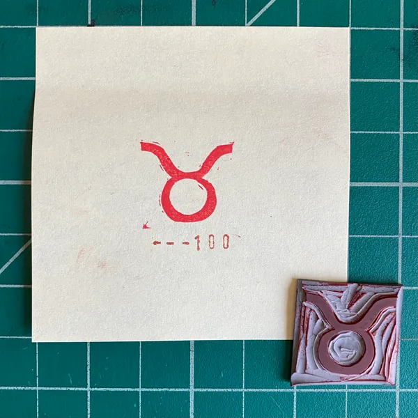 A cream square rests on green grid paper, displaying a red Taurus symbol and dashed lines; to the right is a small, textured square object in maroon and gray tones.