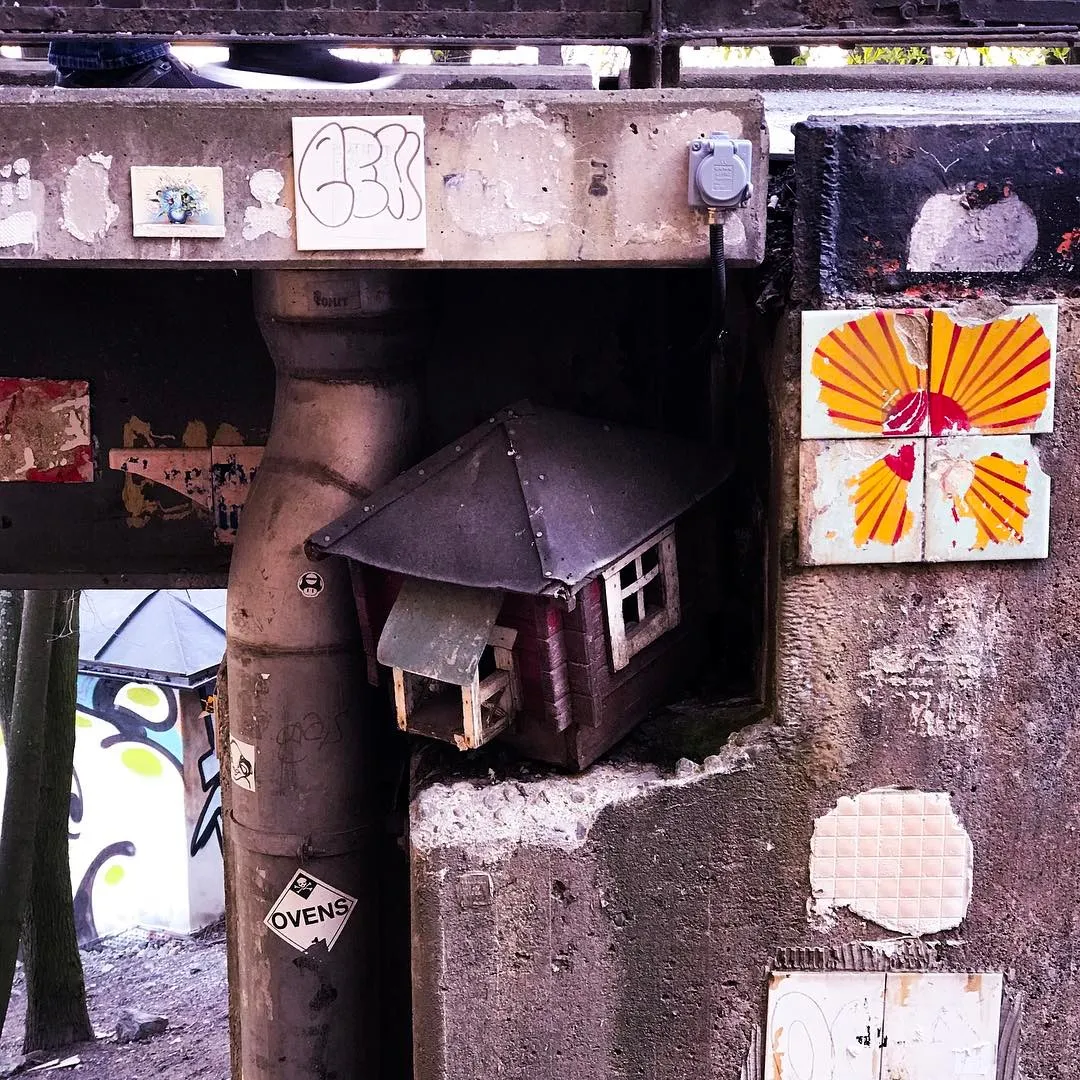 Concrete corner structure with a drainpipe and a small, dark shed, marked by graffiti, peeling paint, and colorful tiles depicting scallop shells.