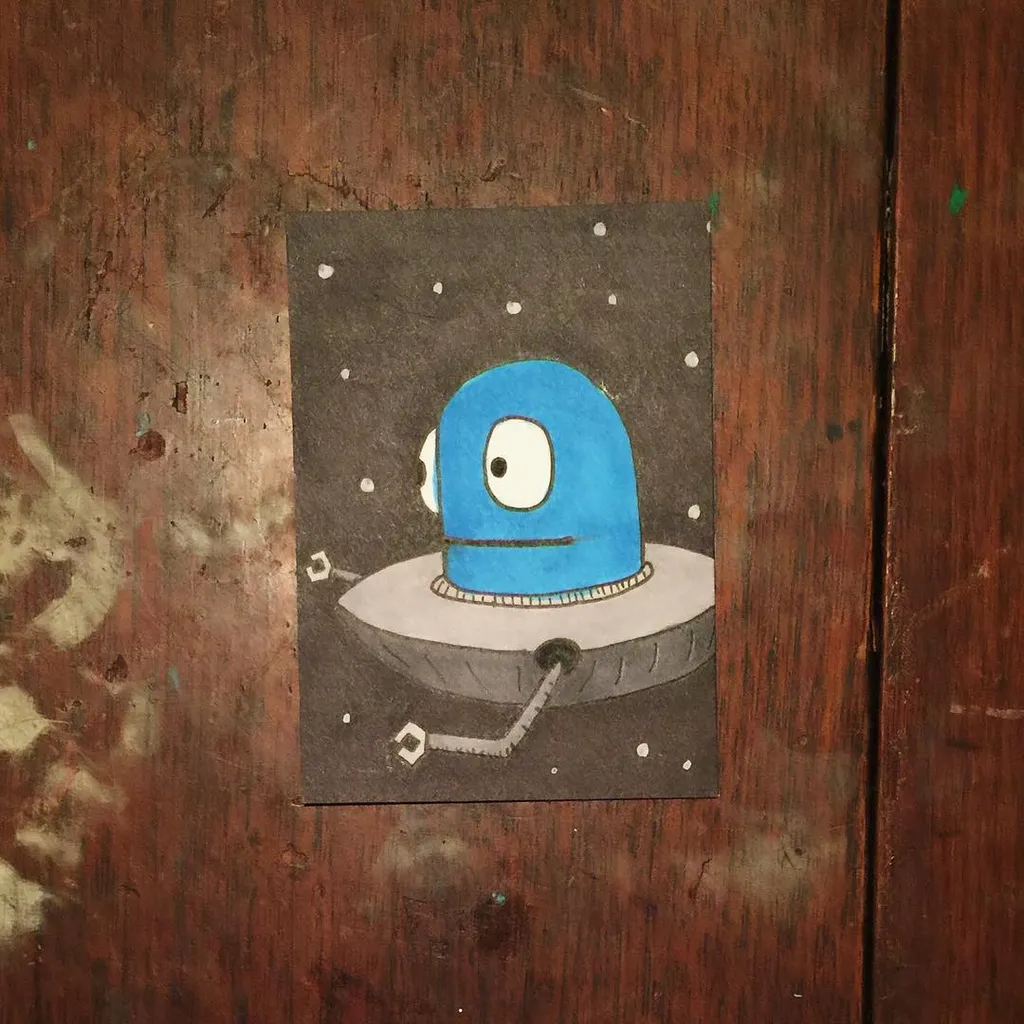 A cartoon blue and gray flying saucer with large eyes floats against a dark background speckled with white dots, mounted on a square piece of paper against dark wood.