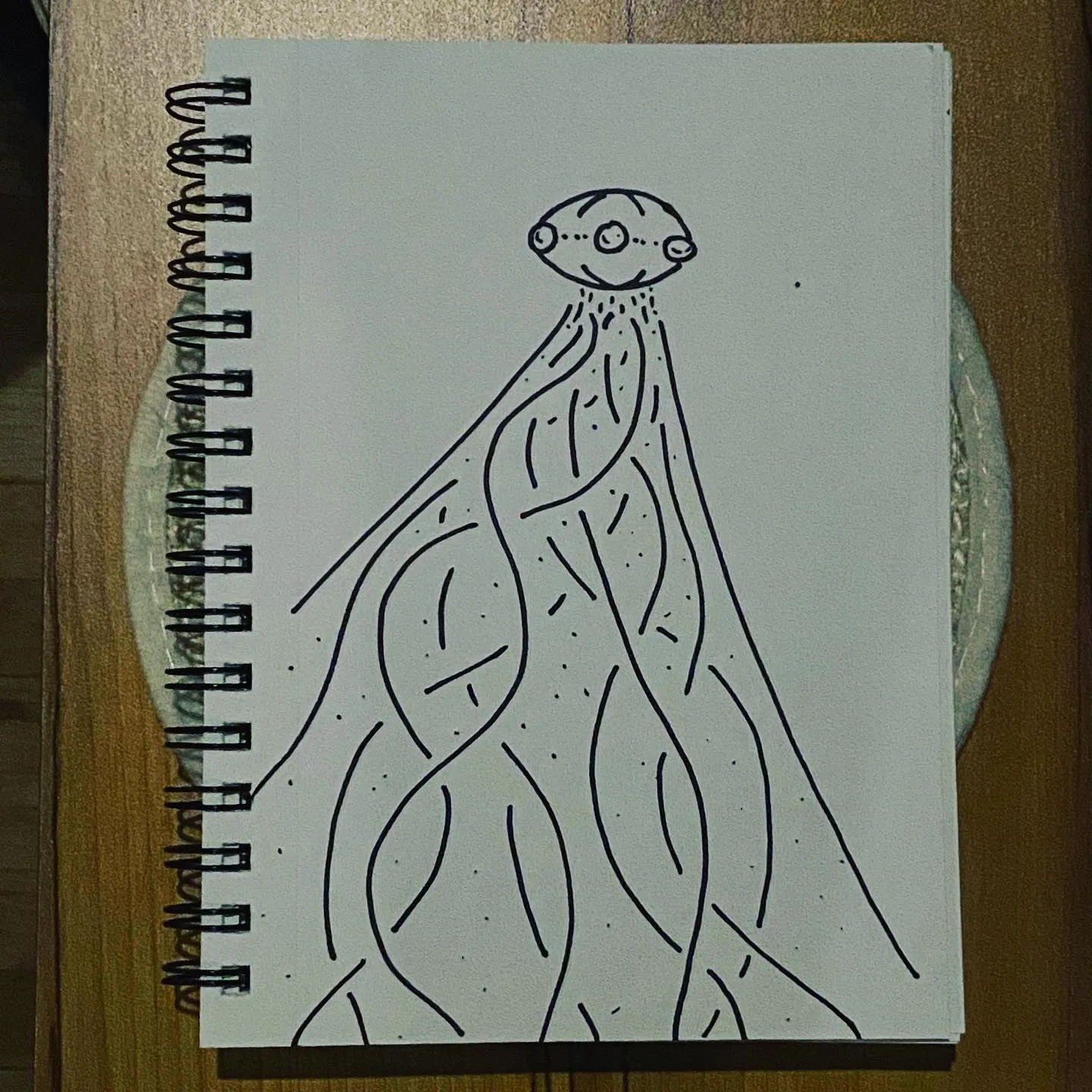 A black ink line drawing on white paper depicts a stylized creature with a round head and multiple flowing, sinuous appendages that cascade downward.