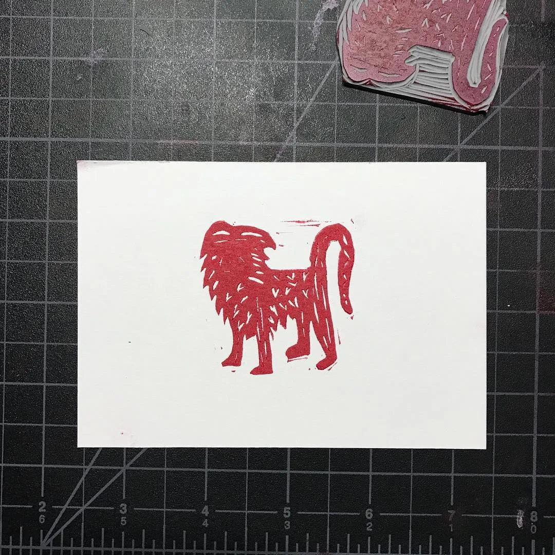 A stylized red creature, rendered with thick vertical lines, stands centered on a white square paper placed on a dark gridded surface.
