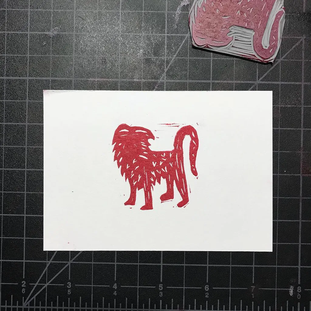 A stylized red creature, rendered with thick vertical lines, stands centered on a white square paper placed on a dark gridded surface.