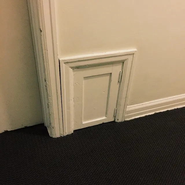 Light beige walls meet a dark carpet at a corner, featuring white painted baseboards and a small, rectangular decorative panel set into the wall.