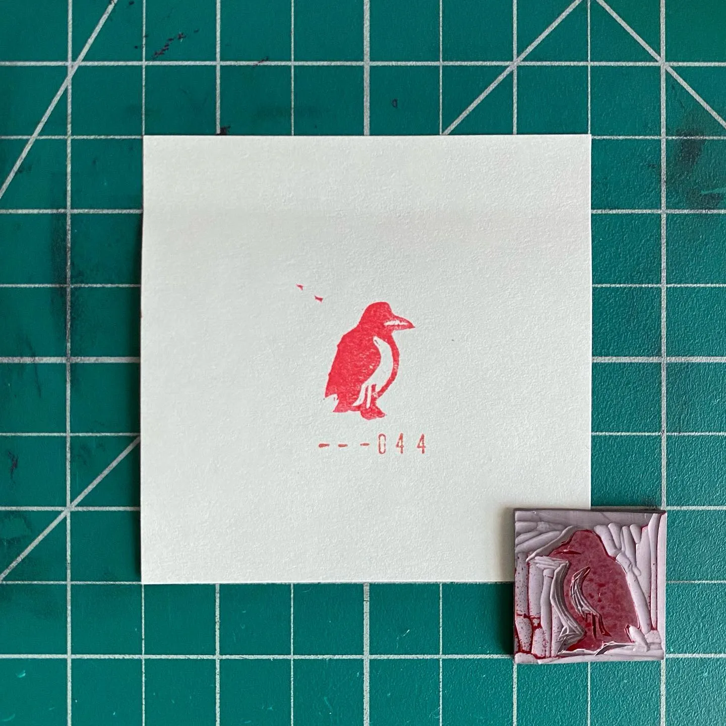 A white card with a stylized red bird and red text ——044 rests on a green gridded surface, beside a small, highly textured piece featuring red and white washes.