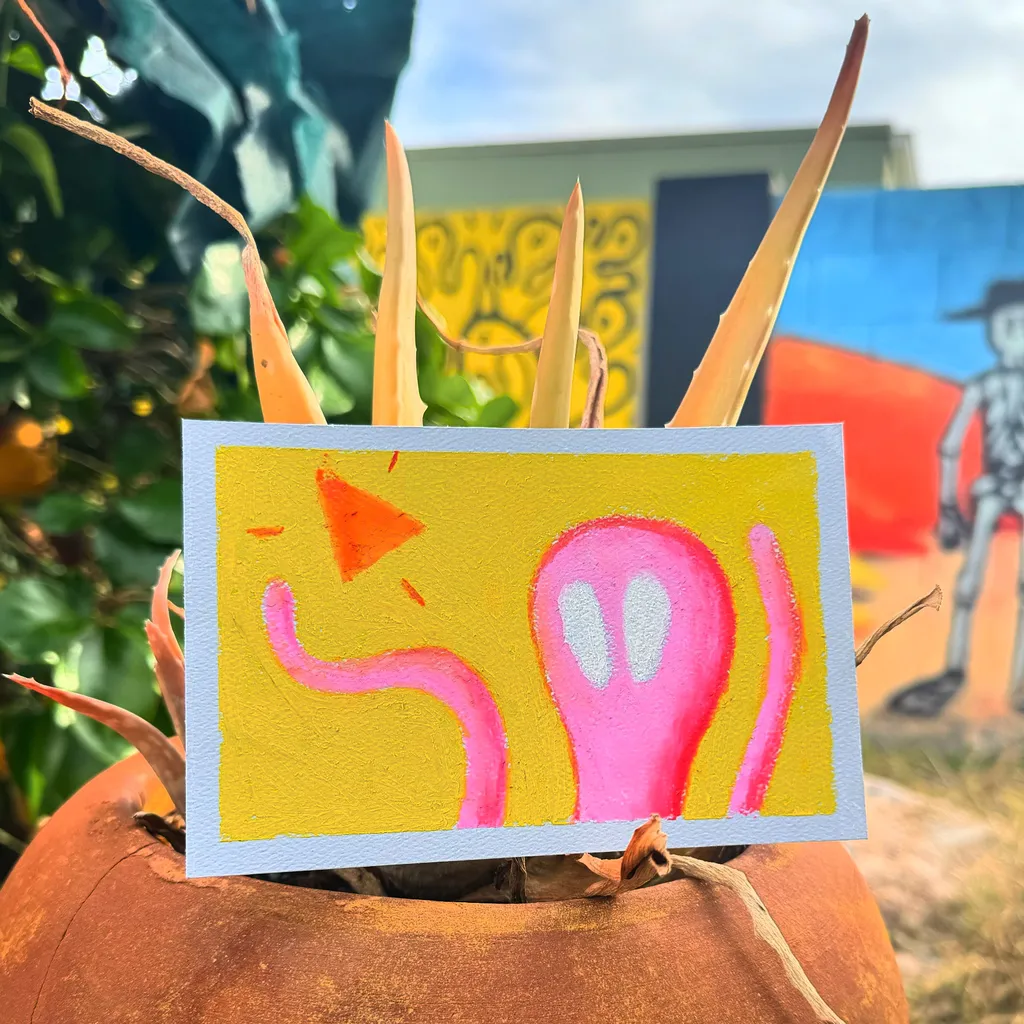 Two yellow artworks featuring abstract pink and orange shapes rest on a textured orange pumpkin, positioned outdoors against a colorful mural.