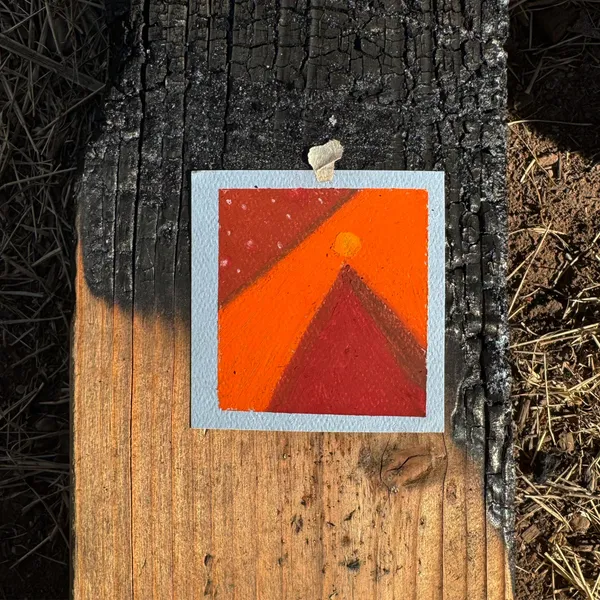 A small, square artwork featuring geometric orange and red shapes with a yellow circle is affixed to a heavily weathered, dark brown wooden post with visible cracks and grain.