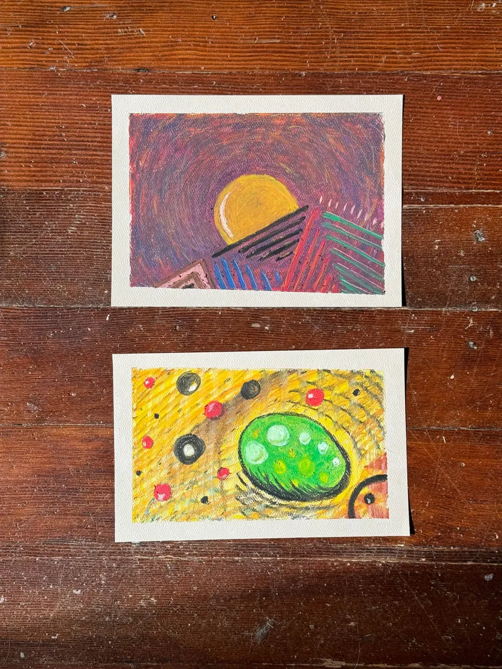 Two abstract paintings rest on dark wood: the top piece shows a yellow circle above red geometric lines, and the bottom piece features a central green oval surrounded by yellow dots and black marks.