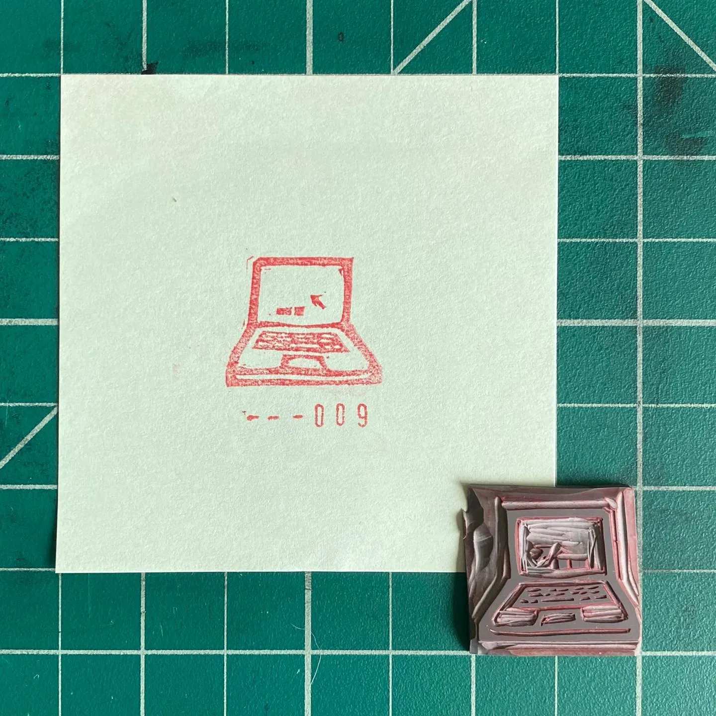 A light paper on a teal grid displays a red outline drawing of an open laptop and the number 600-009, with a matching reddish-brown object positioned below the paper.
